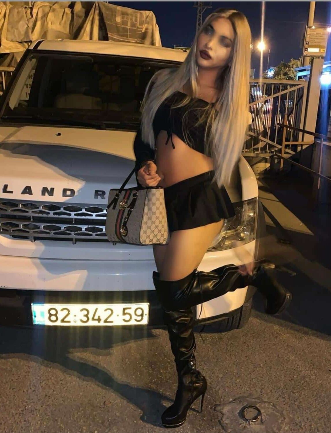 WhatsApp-Image-2020-12-28-at-12.19.48 אישה עם שיער בלונדיני ארוך עומדת ליד רכב לבן בלילה. היא לובשת חצאית שחורה קצרה, חולצה שחורה עם כתפיים חשופות ומגפיים שחורים גבוהים. היא מחזיקה תיק מעוצב ביד אחת ומרימה את רגל ימין.
