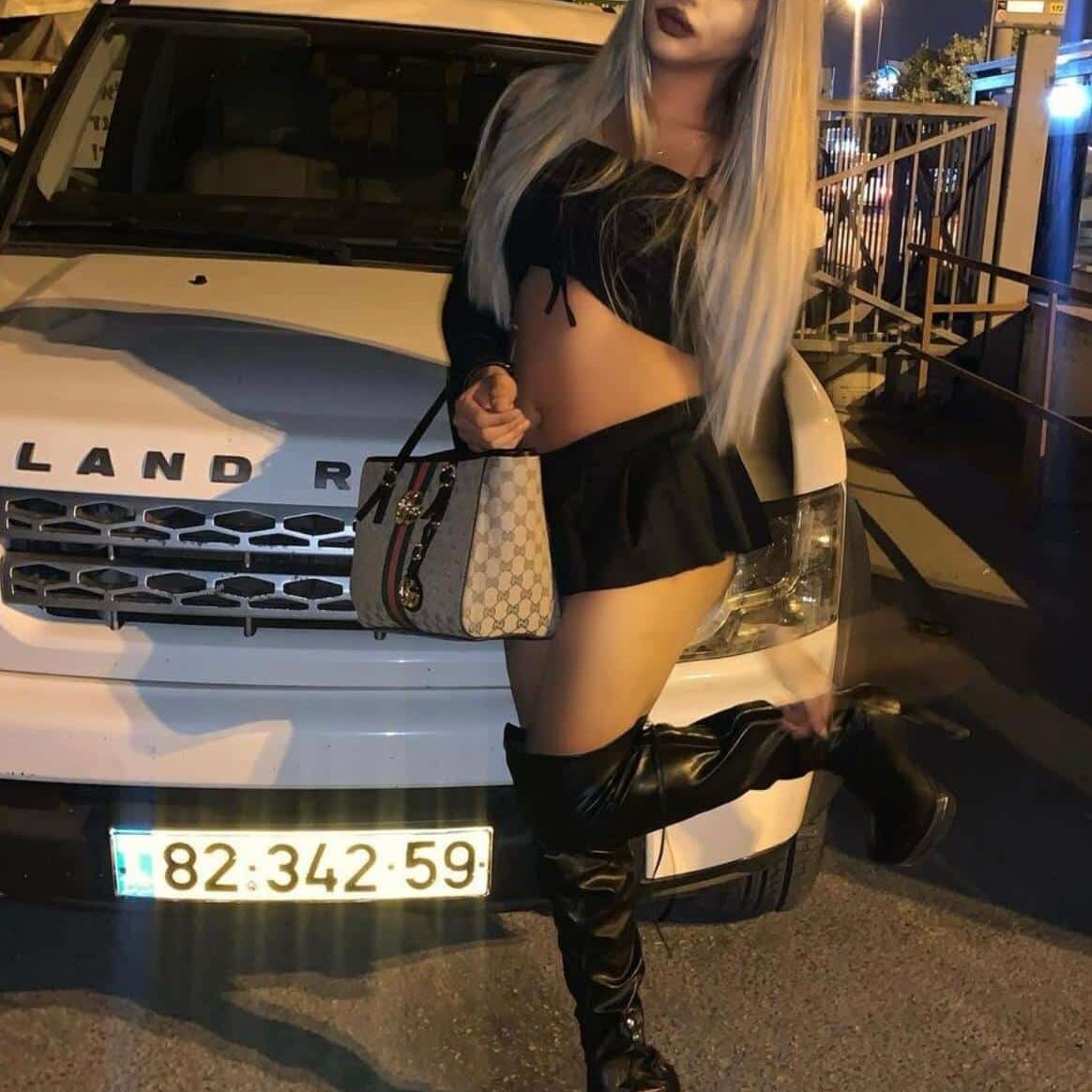 אישה עם שיער בלונדיני ארוך עומדת ליד רכב לבן בלילה. היא לובשת חצאית שחורה קצרה, חולצה שחורה עם כתפיים חשופות ומגפיים שחורים גבוהים. היא מחזיקה תיק מעוצב ביד אחת ומרימה את רגל ימין.