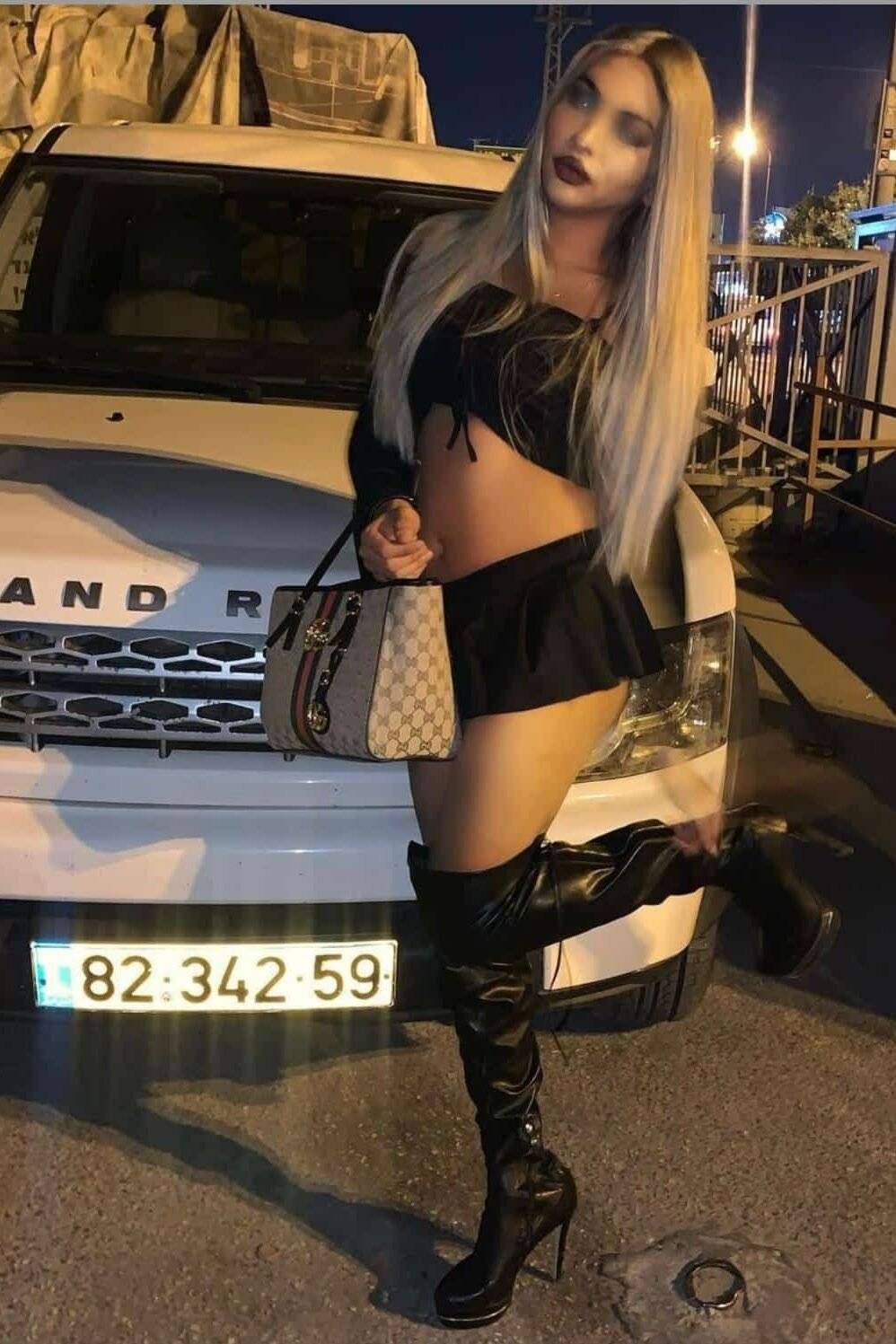 WhatsApp-Image-2020-12-28-at-12.19.48 אישה עם שיער בלונדיני ארוך עומדת ליד רכב לבן בלילה. היא לובשת חצאית שחורה קצרה, חולצה שחורה עם כתפיים חשופות ומגפיים שחורים גבוהים. היא מחזיקה תיק מעוצב ביד אחת ומרימה את רגל ימין.