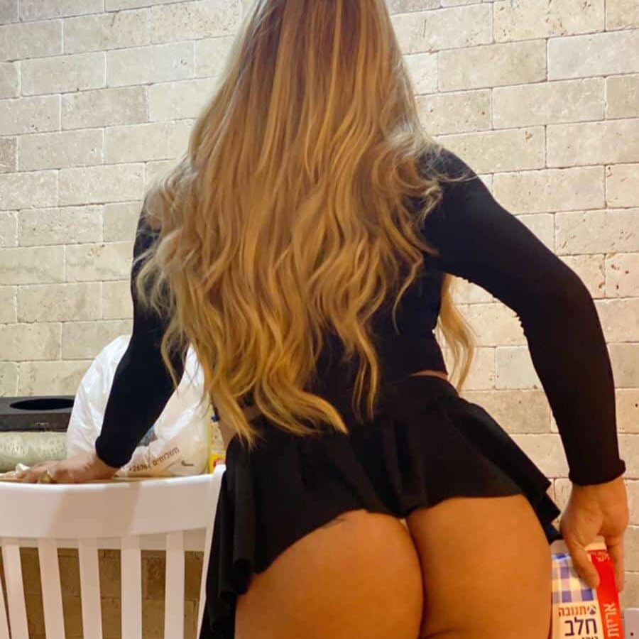 אישה עם שיער ארוך לובשת חולצה שחורה קצרה ומגפיים גבוהים, אוחזת במיכל חלב במטבח.