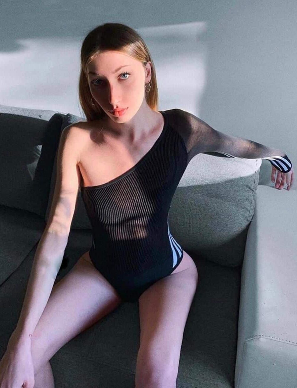 אישה יושבת על ספה ומשתזפת באור שמש, לבושה בבגד גוף שחור עם כתף אחת חשופה.