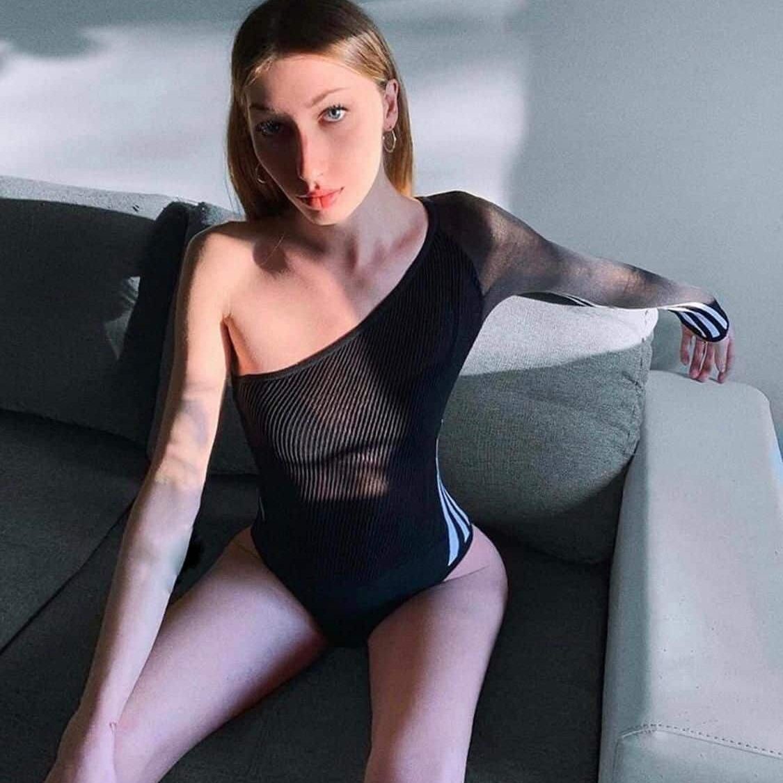 אישה יושבת על ספה ומשתזפת באור שמש, לבושה בבגד גוף שחור עם כתף אחת חשופה.