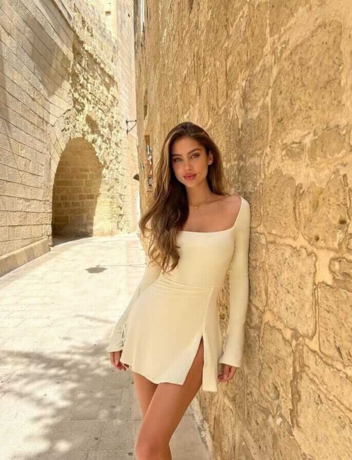 אישה לובשת שמלה לבנה צרה עם שרוולים ארוכים, עומדת ליד קיר אבן ברחוב עתיק ותאורה טבעית חזקה.