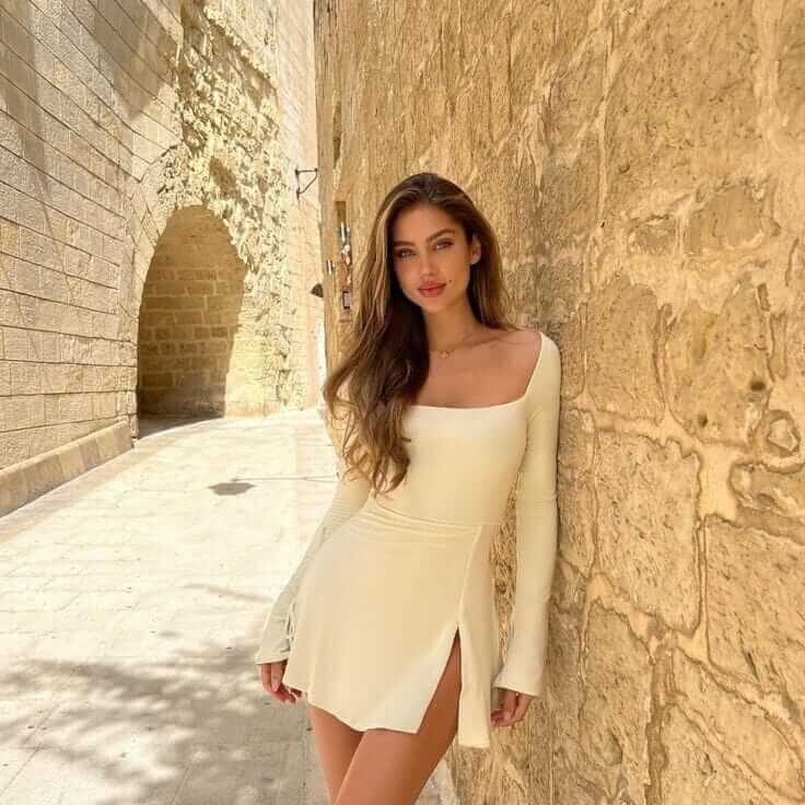 אישה לובשת שמלה לבנה צרה עם שרוולים ארוכים, עומדת ליד קיר אבן ברחוב עתיק ותאורה טבעית חזקה.