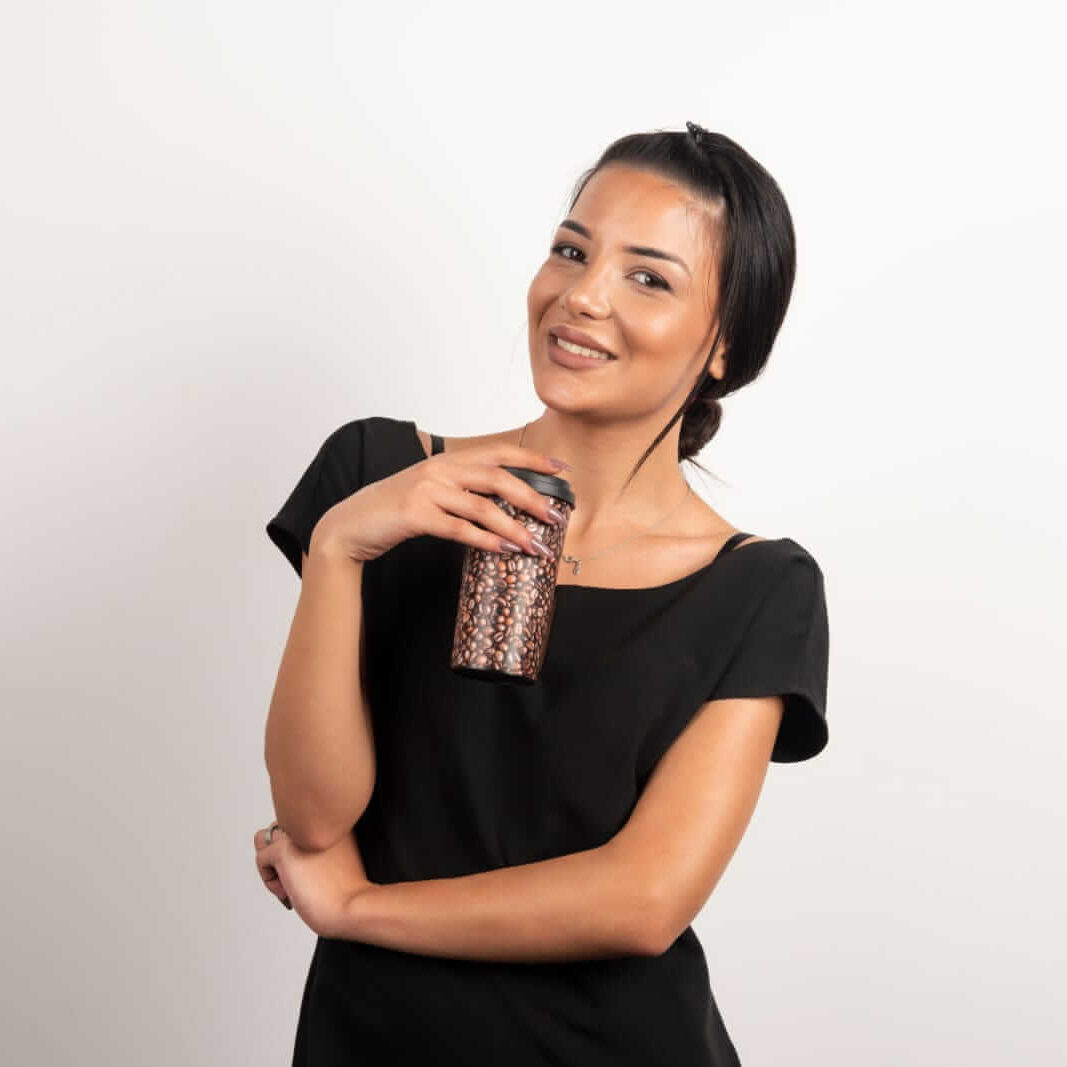 אישה לבושה בשחור מחזיקה כוס לשתייה עם דוגמאות חומות, על רקע לבן.