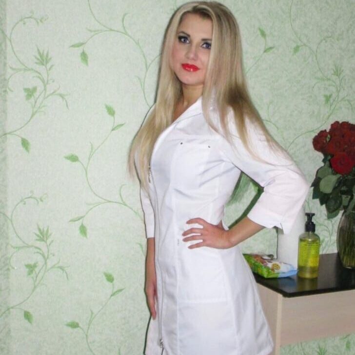 אישה בלונדינית לובשת חלוק לבן עומדת ליד שולחן עם אגרטל פרחים אדומים ובקבוק סבון.