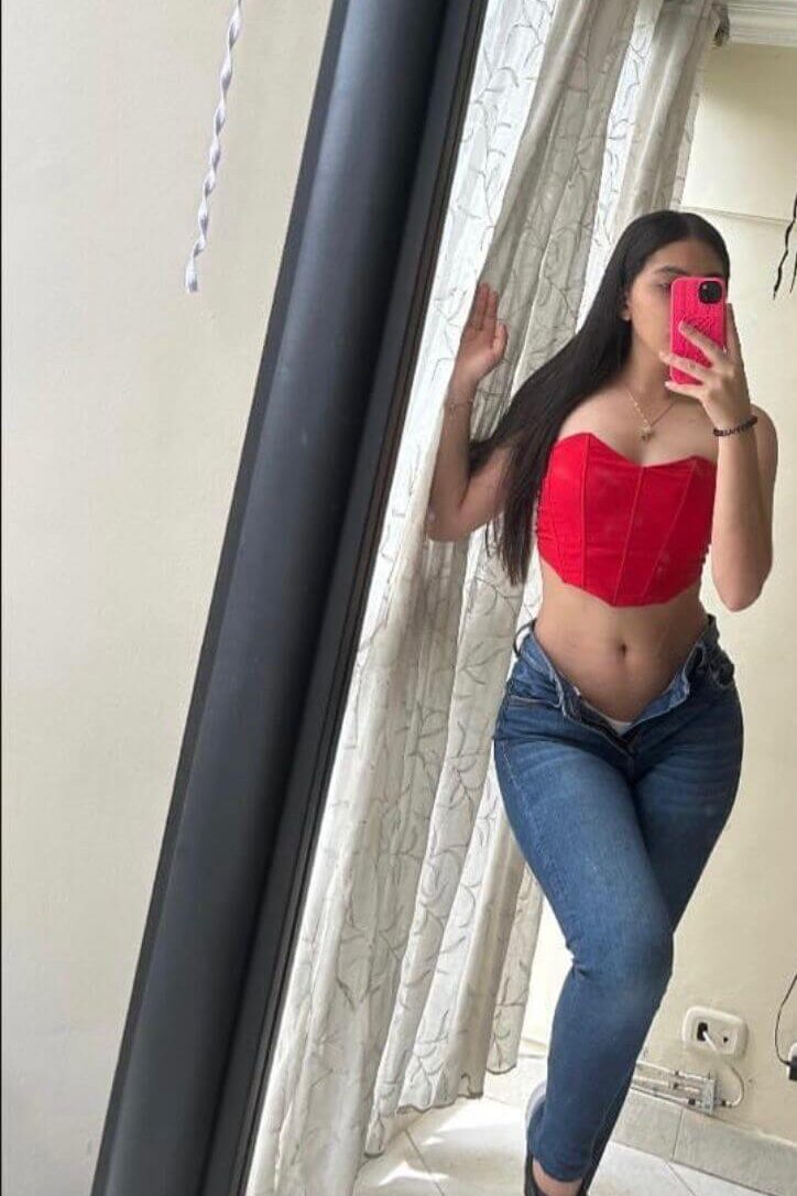 תל-אביב-ישראלית-צברית-24 אישה צעירה מצלמת סלפי במראה, לובשת ג'ינס וחולצה אדומה, מחזיקה טלפון עם כיסוי ורוד.