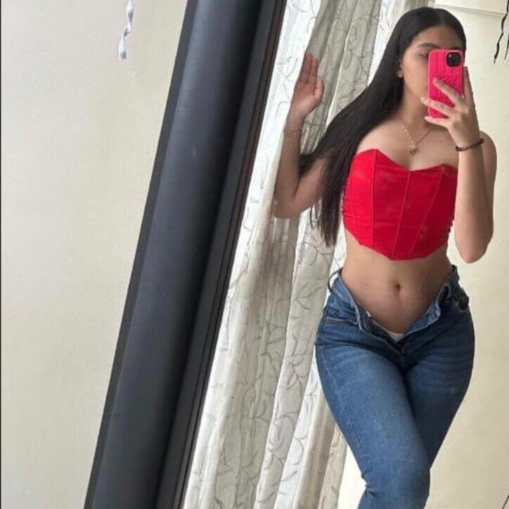 אישה צעירה מצלמת סלפי במראה, לובשת ג'ינס וחולצה אדומה, מחזיקה טלפון עם כיסוי ורוד.