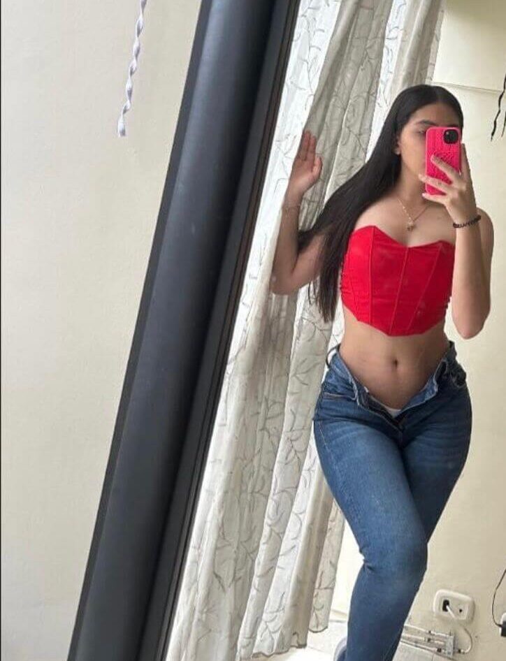 תל-אביב-ישראלית-צברית-24 אישה צעירה מצלמת סלפי במראה, לובשת ג'ינס וחולצה אדומה, מחזיקה טלפון עם כיסוי ורוד.