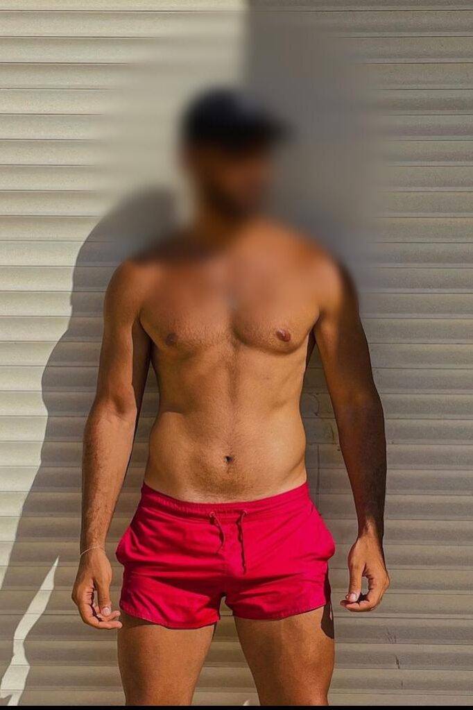 WhatsApp-Image-2023-01-09-at-14.16.53 גבר עומד מול קיר עם תריסים לבנים, לובש מכנסיים קצרים אדומים וכובע, הגוף מטושטש בפנים ובפרטים מזהים.