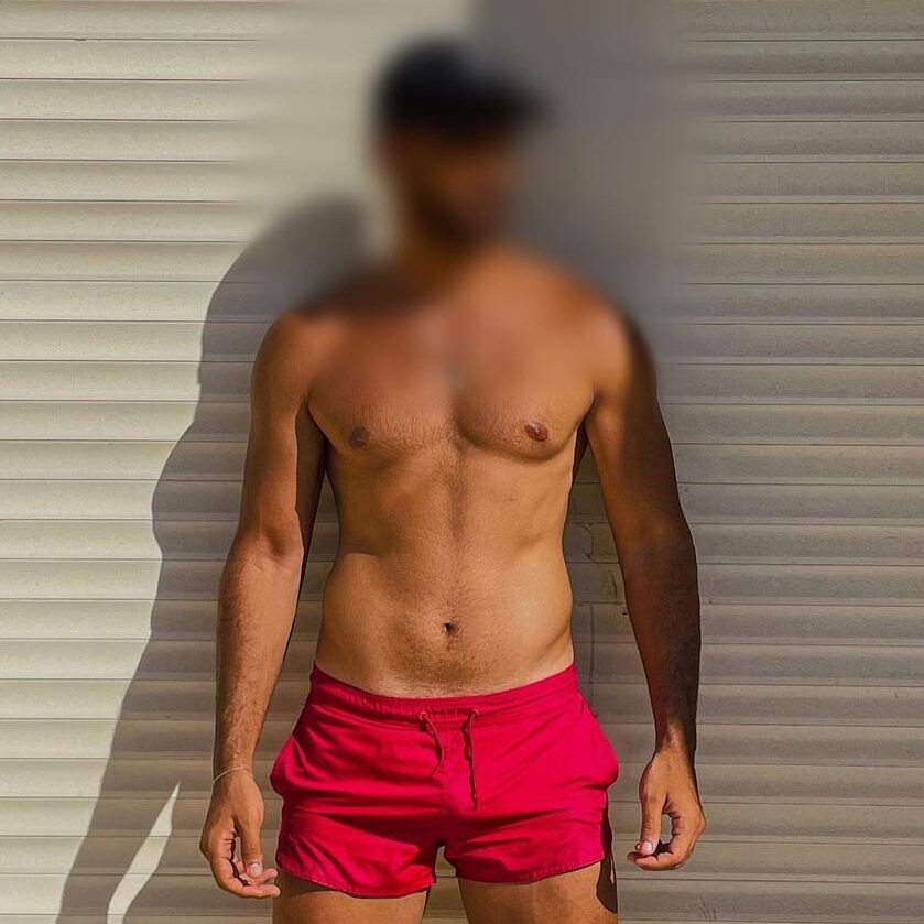 גבר עומד מול קיר עם תריסים לבנים, לובש מכנסיים קצרים אדומים וכובע, הגוף מטושטש בפנים ובפרטים מזהים.