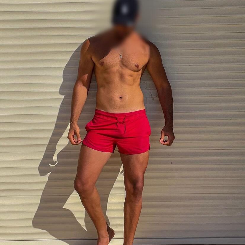 גבר לובש מכנסיים קצרים אדומים וצמיד כסוף עומד בצל על רקע תריס.