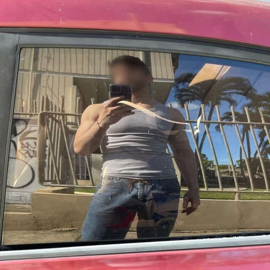 דמות משתקפת בחלון מכונית אדומה עם אדן זהוב, לובשת גופייה אפורה ומכנסי ג'ינס, על רקע מבנה עם סורגים ועצים.