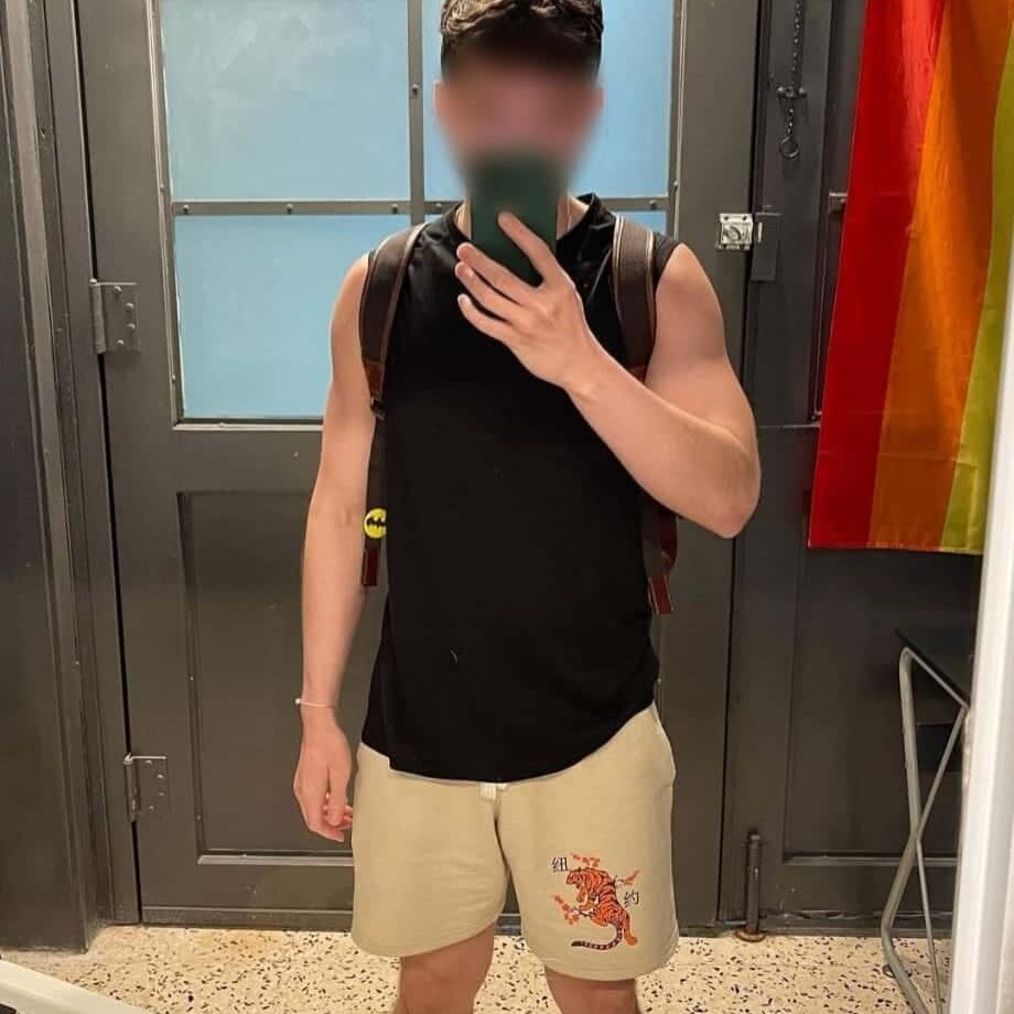 גבר מצולם מול מראה, לובש גופייה שחורה ומכנסיים קצרים בצבע בז' עם ציור נמר עליהם. דגל בצבעי גאווה תלוי על הקיר.