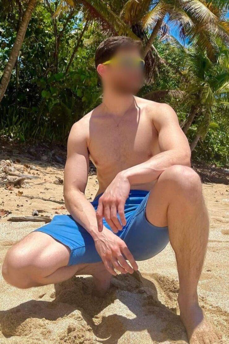 WhatsApp-Image-2023-05-22-at-09.08.46-2 אדם ללא חולצה עם מכנסיים קצרות כחולות, כורע על חול בחוף ים עם עצי דקל ברקע.