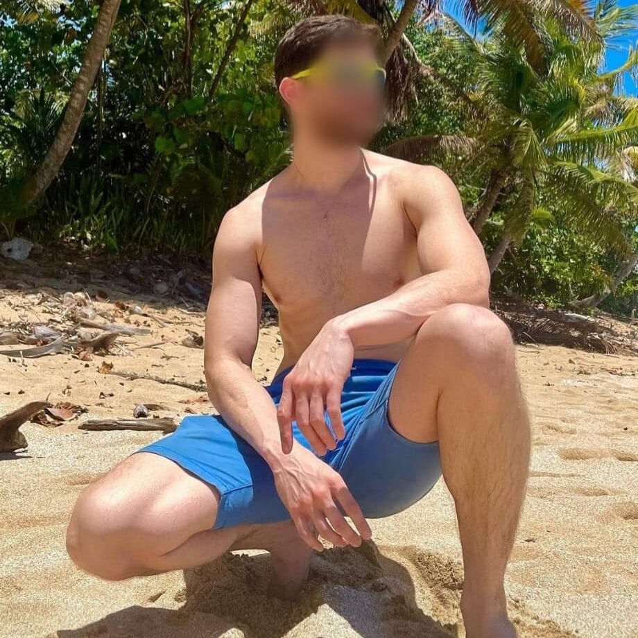 אדם ללא חולצה עם מכנסיים קצרות כחולות, כורע על חול בחוף ים עם עצי דקל ברקע.