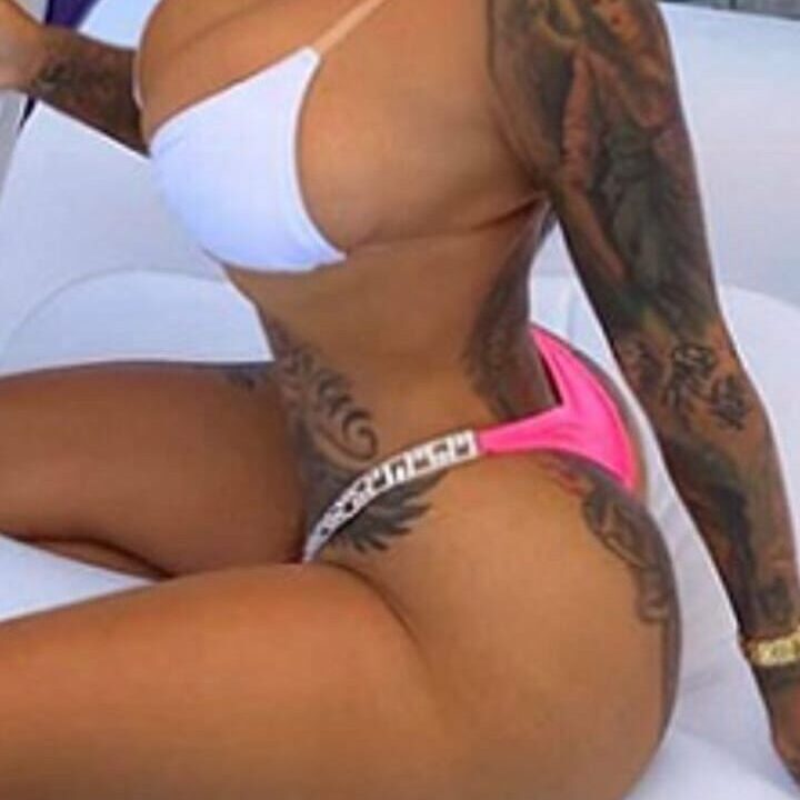 אישה לובשת בגד ים לבן וורוד יושבת על מצוף, עם קעקועים בולטים על זרועה וגופה.