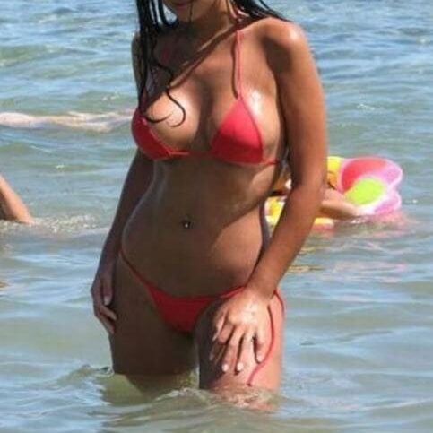 אישה בלבוש ביקיני אדום עומדת במים הרדודים על חוף הים, כאשר גלים קטנים נשברים ברקע.