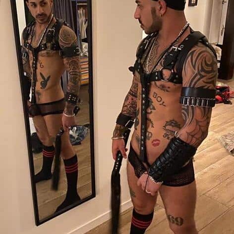 אדם עומד מול מראה, לבוש בביגוד עור, עם קעקועים על גופו ומחזיק ביד חפץ המשמש פורק או מגלב, בתוך חדר מעוצב בסגנון מודרני.