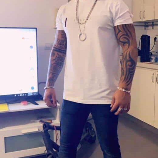 גבר קירח לבוש בחולצת טריקו לבנה ומכנסי ג'ינס כהים, עומד בחדר ליד טלוויזיה ומוצרי מדיה, עם קעקועים על ידיו השמאלית והימנית.