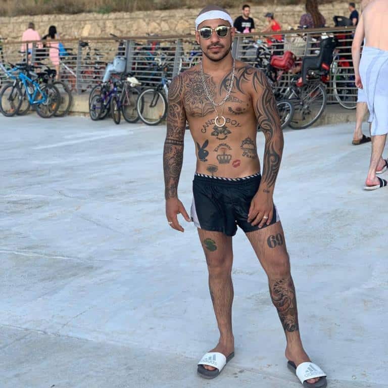 אדם מקועקע לבוש מכנסיים קצרים וכפכפים עומד על טיילת עם אופניים ברקע.