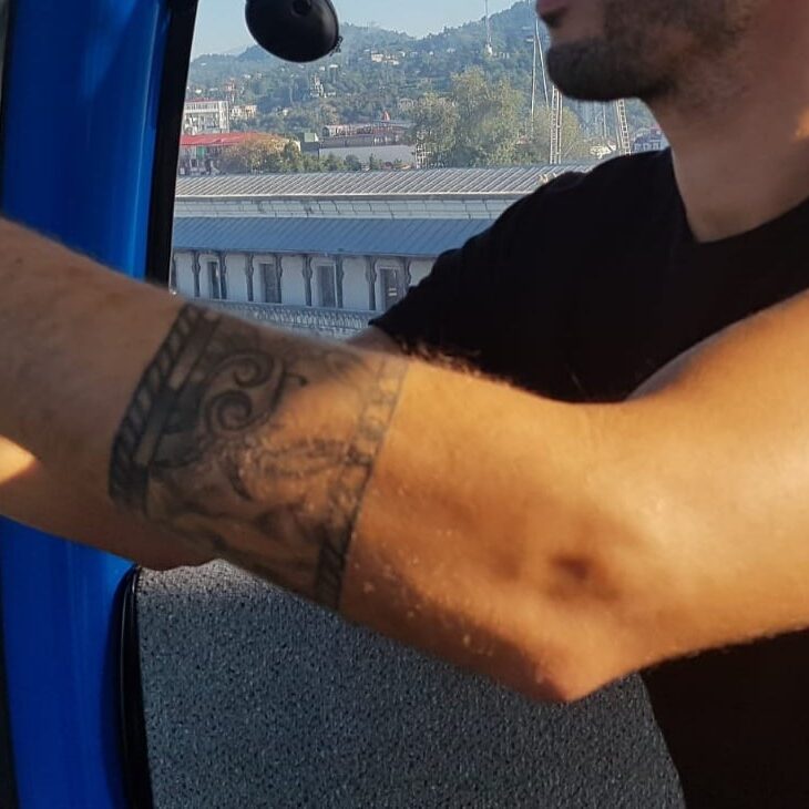 גבר לובש חולצה שחורה עם קעקוע על הזרוע שלו יושב במתקן תחבורה עילי כאשר בניינים ונוף עירוני רואים בחלון מאחור.