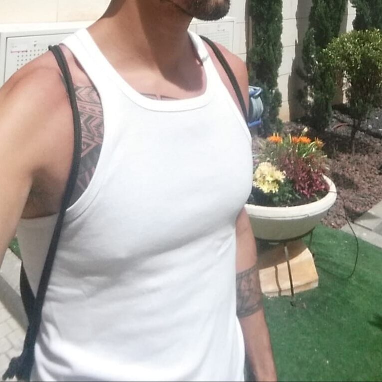 גבר לובש גופייה לבנה עם קעקועים על הכתפיים מדלג בגינה מעוצבת עם עציצים ופרחים.