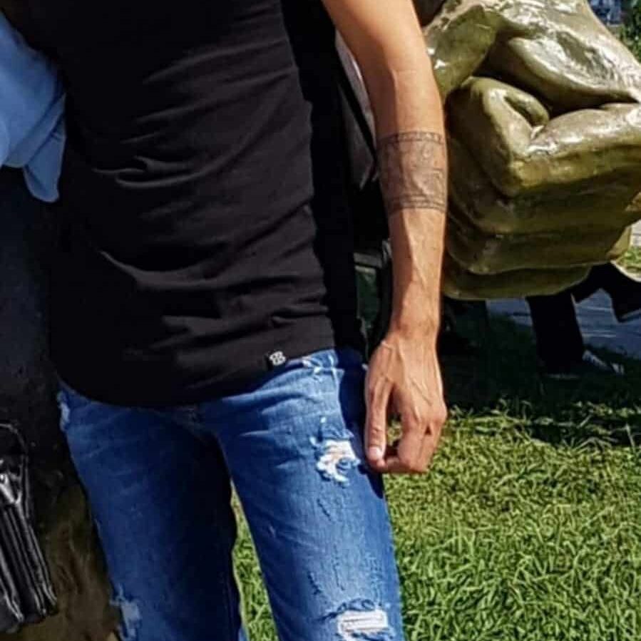 אדם בפאה העליונה לבוש בחולצה שחורה וג'ינס קרועים, עומד ליד פסל של יד גדולה באוויר פתוח.