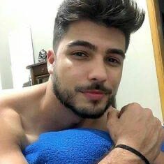 גבר עם שיער כהה וזקן מטופח מחייך כשהוא נשען על כרית בצבע כחול.