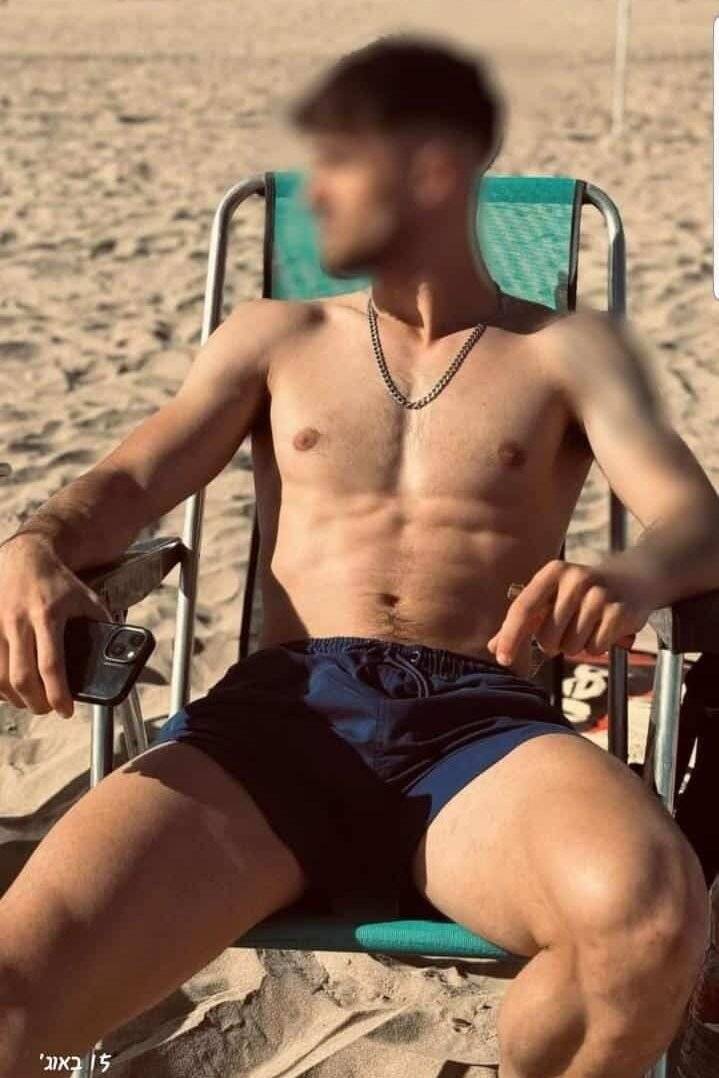 WhatsApp-Image-2024-08-21-at-15.32.38 גבר יושב על כיסא חוף בבגד ים ללא חולצה, מחזיק טלפון בידו, על חוף חול.
