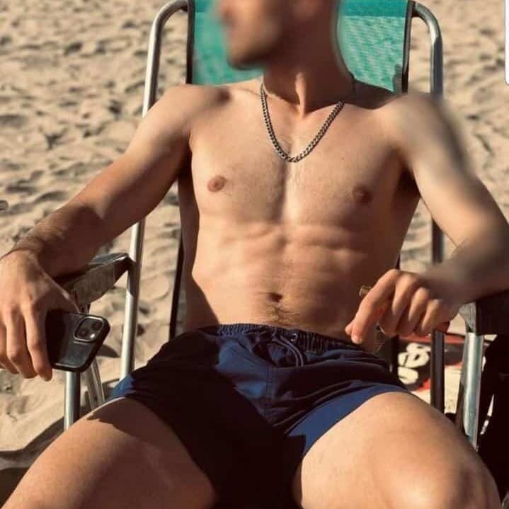 גבר יושב על כיסא חוף בבגד ים ללא חולצה, מחזיק טלפון בידו, על חוף חול.