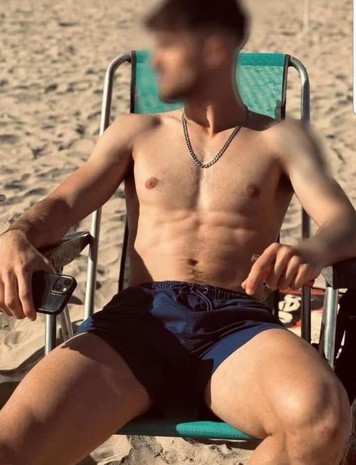 גבר יושב על כיסא חוף בבגד ים ללא חולצה, מחזיק טלפון בידו, על חוף חול.