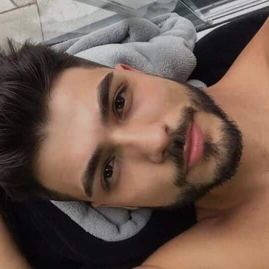 גבר עם שיער מבולגן וזקן קל שוכב על מצע רך, מביט למצלמה.