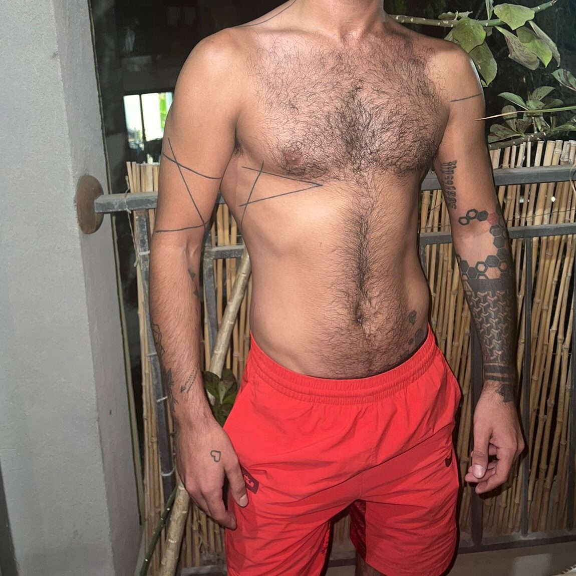 גבר עירום חזה עם קעקועים גאומטריים על זרועותיו ולבוש מכנסיים קצרים כתומים עומד ליד גדר במרפסת.