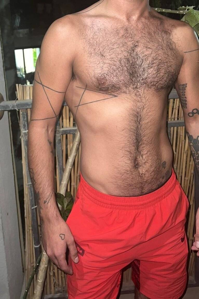 WhatsApp-Image-2024-12-19-at-11.16.05-2 גבר עירום חזה עם קעקועים גאומטריים על זרועותיו ולבוש מכנסיים קצרים כתומים עומד ליד גדר במרפסת.