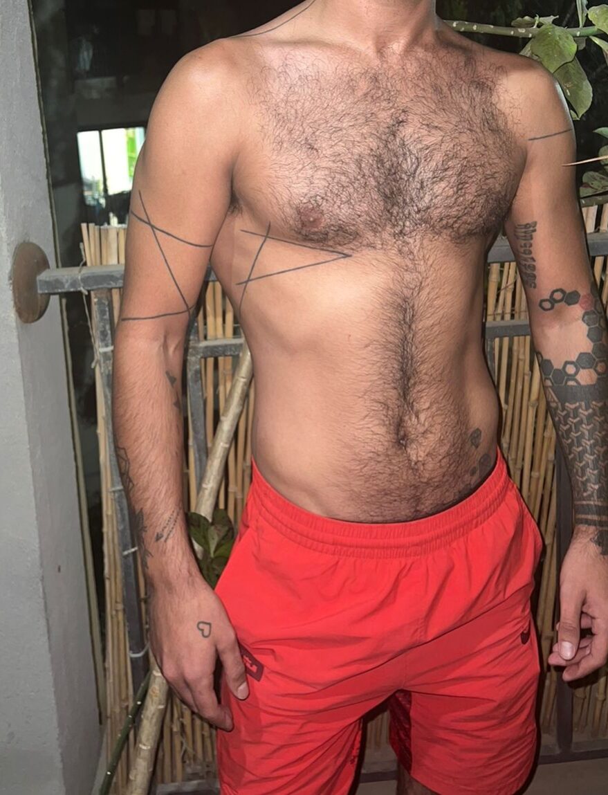 WhatsApp-Image-2024-12-19-at-11.16.05-2 גבר עירום חזה עם קעקועים גאומטריים על זרועותיו ולבוש מכנסיים קצרים כתומים עומד ליד גדר במרפסת.