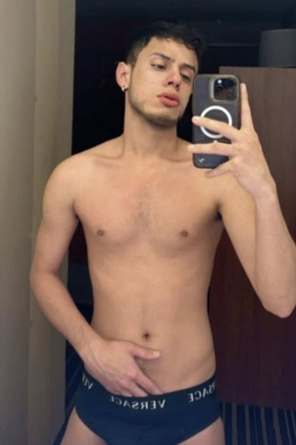 WhatsApp-Image-2024-12-25-at-06.56.14 גבר צעיר מצלם סלפי במראה כשהוא לובש תחתוני ורסאצ'ה שחורים בחדר.