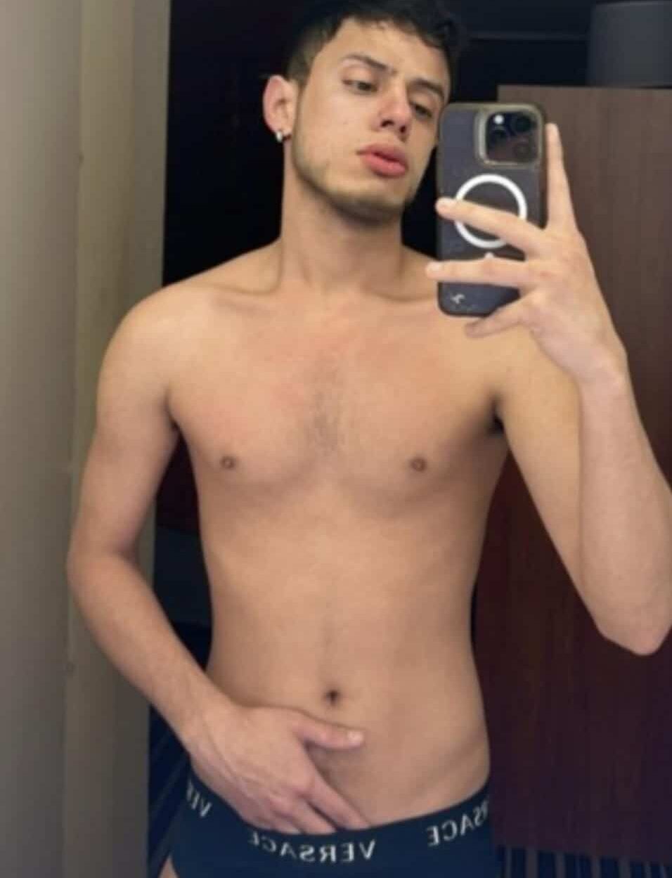 WhatsApp-Image-2024-12-25-at-06.56.14 גבר צעיר מצלם סלפי במראה כשהוא לובש תחתוני ורסאצ'ה שחורים בחדר.