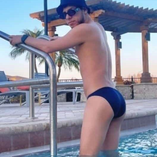 אדם עומד בבריכת שחייה, ליד סולם, לובש בגדי ים כהים, כובע משקפי שמש, וקיים ברקע רהיטי גן ועצים.