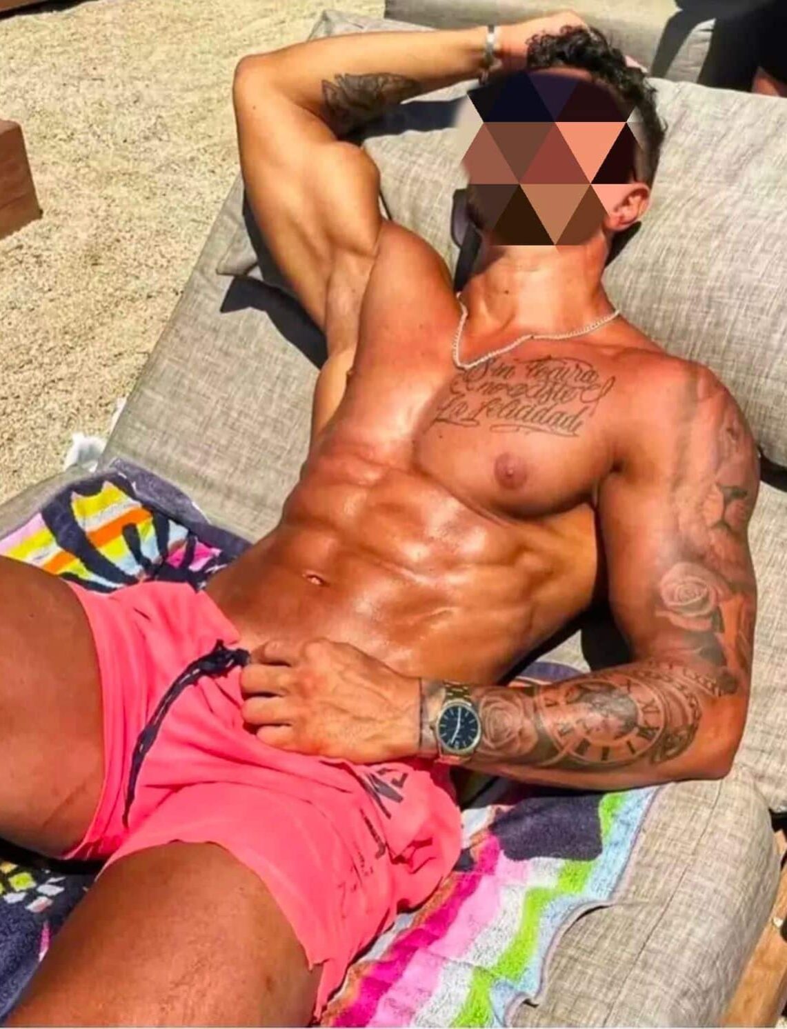WhatsApp-Image-2025-01-13-at-18.16.44 גבר שוכב על מיטת חוף בבגד ים ורוד, עם קעקועים על הזרוע וכתפיים, ושרשרת על הצוואר.