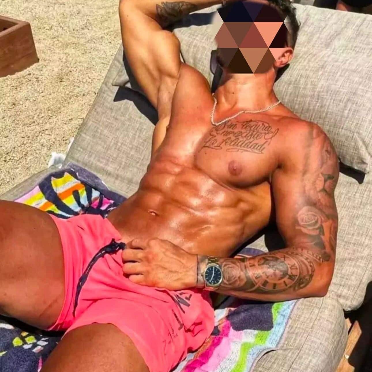 גבר שוכב על מיטת חוף בבגד ים ורוד, עם קעקועים על הזרוע וכתפיים, ושרשרת על הצוואר.