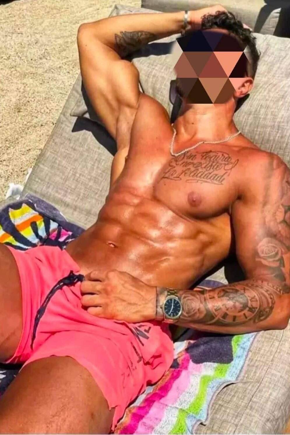 WhatsApp-Image-2025-01-13-at-18.16.44 גבר שוכב על מיטת חוף בבגד ים ורוד, עם קעקועים על הזרוע וכתפיים, ושרשרת על הצוואר.