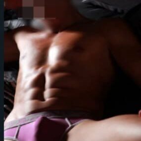 גבר עם פנים מטושטשות שוכב על הגב לובש תחתונים סגולים, שרירי גופו מודגשים בתאורה עמומה.