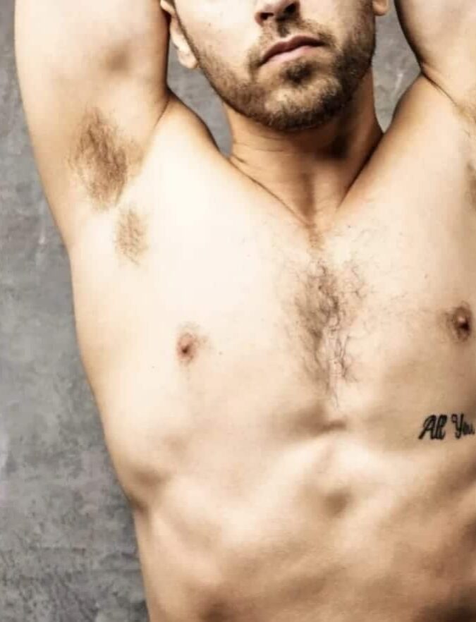 WhatsApp-Image-2025-01-26-at-13.46.10-2 פלג גוף עליון של גבר חשוף חזה עם כתובת קעקוע על בית החזה, על רקע קיר אפור.