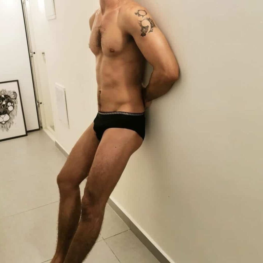 גבר עומד במסדרון כשהוא לובש תחתונים בלבד, עם קעקוע על הזרוע ובחדר רואים תמונת אריה על הרצפה.