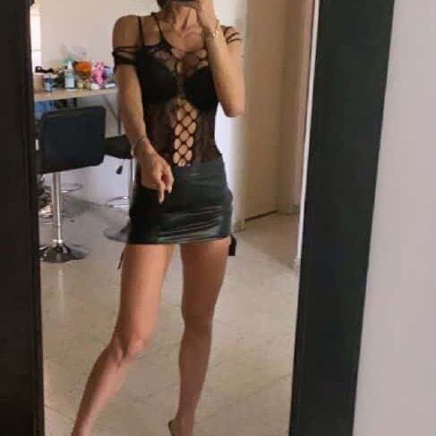 אישה מצלמת את עצמה במראה, לבושה בבגד עליון בצבע שחור עם דוגמא פתוחה וחצאית קצרה שחורה.